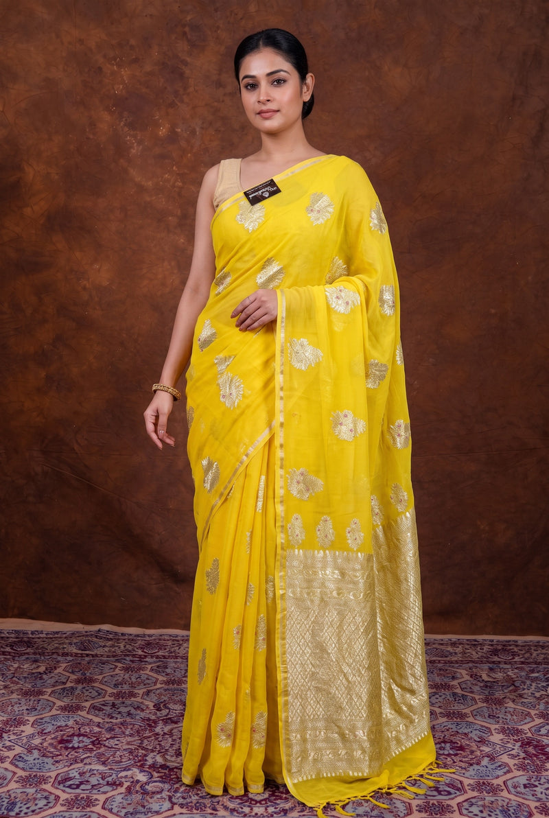 A woman modeling a bright yellow saree with metallic gold floral motifs and a wide gold border, posed against a textured brown backdrop on a patterned rug.