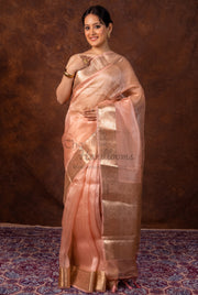 A woman modeling a peach-pink and gold Banarasi silk saree against a textured brown backdrop.